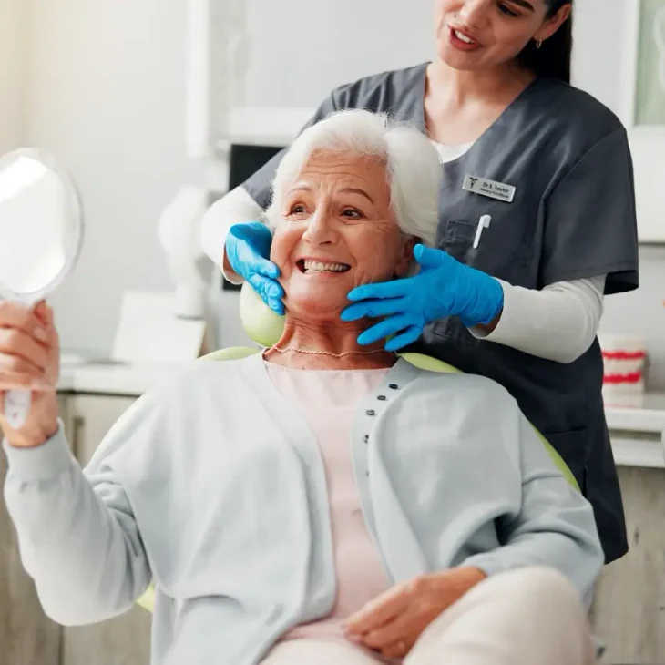 A patient smiling at her reflection in a hand mirror while her hygienist steadies it