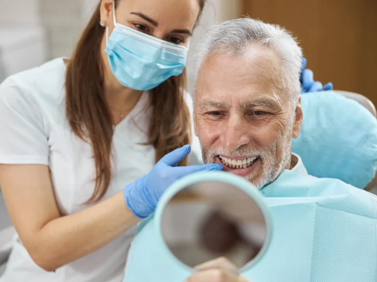 A senior man smiling at his restored smile in a hand mirror held by his hygienist