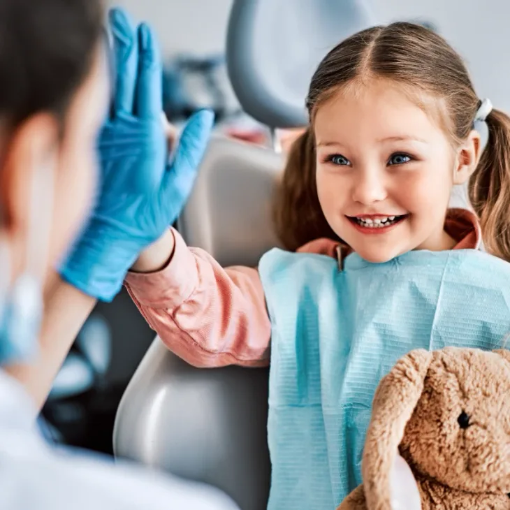 A young girl high-fiving her dentist from the chair, holding a teddy bear